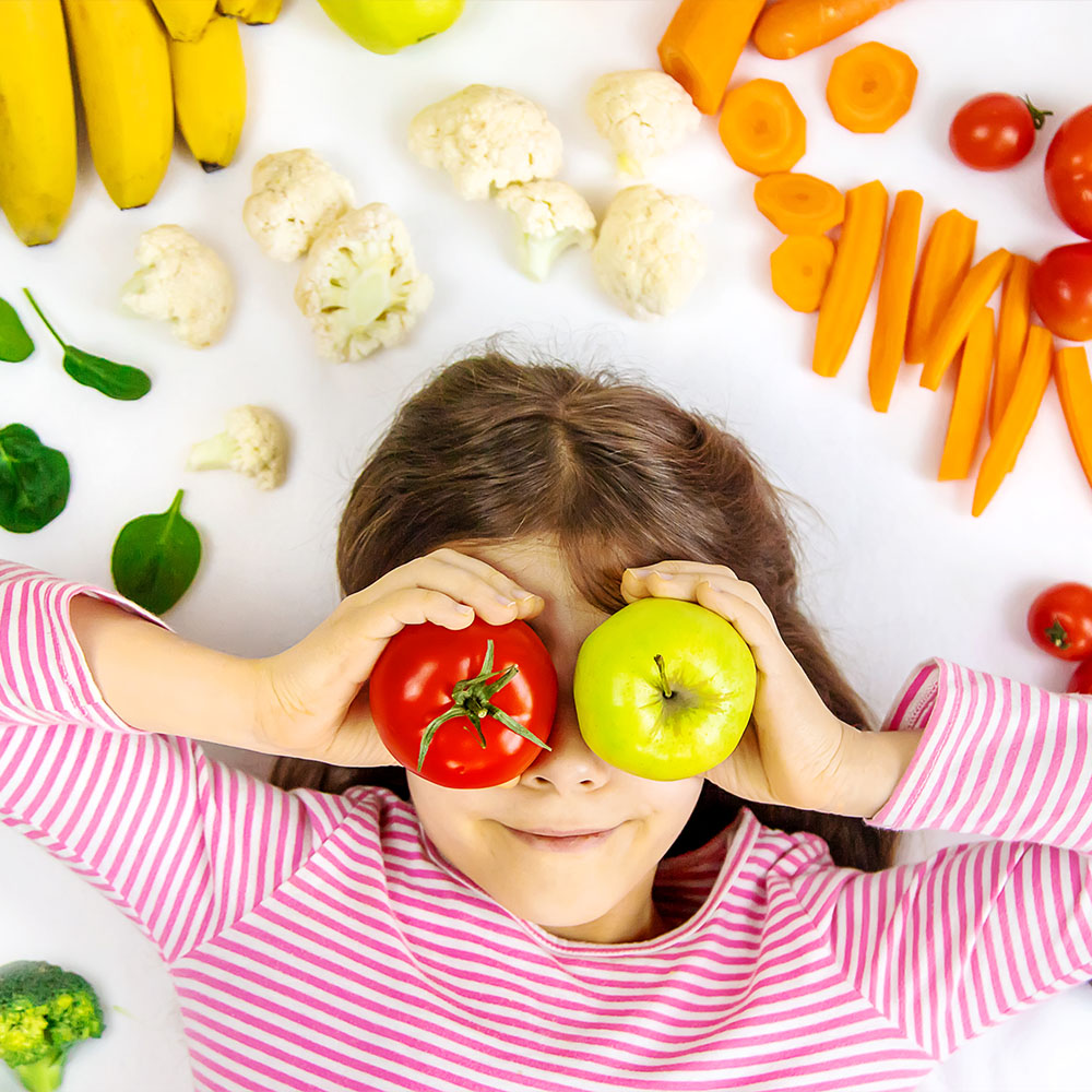 Na imagem, uma criança cercada por frutas e legumes, brinca com um tomate e uma maça verda à frente dos olhos