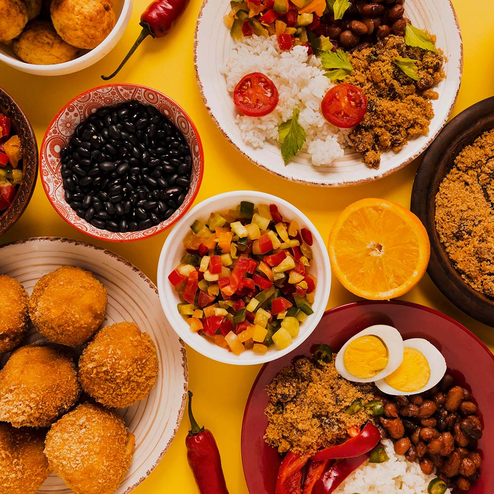 Na imagem aparecem diversos alimentos comuns da culinária e cultura brasileiras, como feijão, farofa, preparos com frutas e legumes