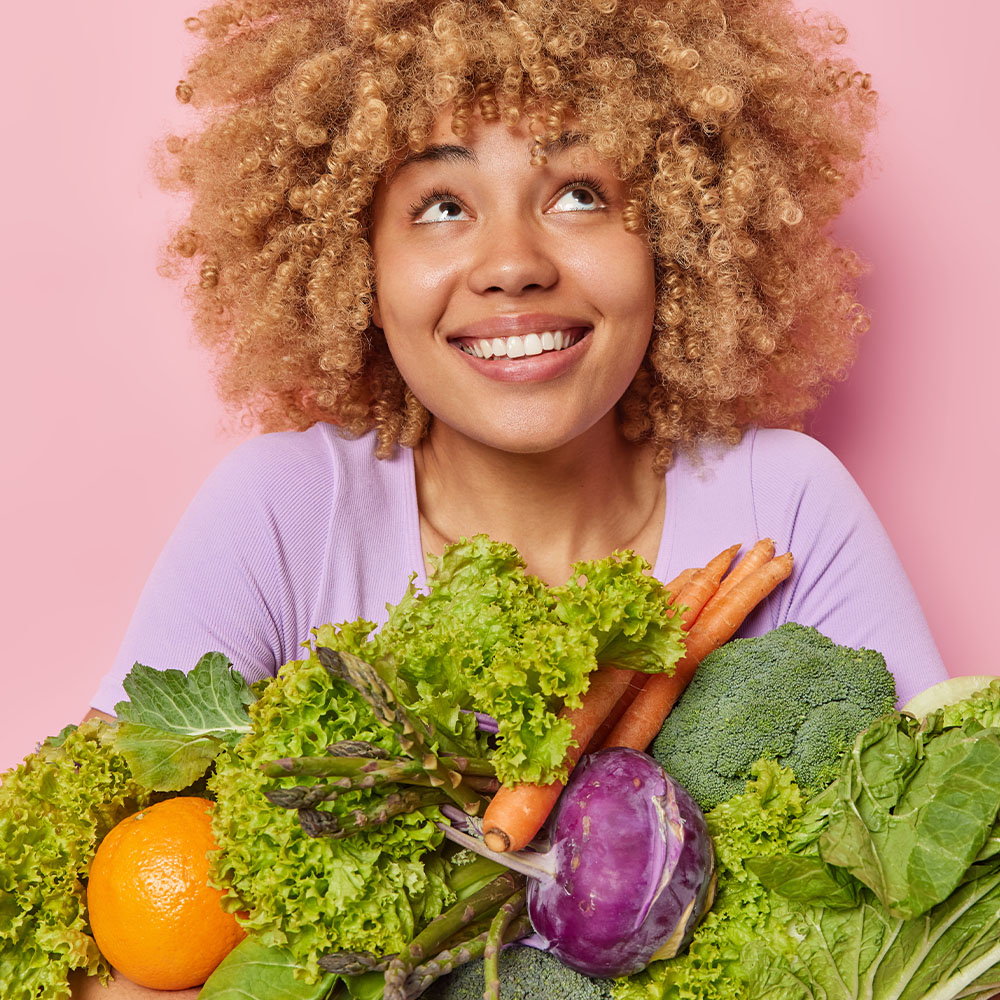 Na imagem, uma mulher sorri rodeada por vegetais e legumes