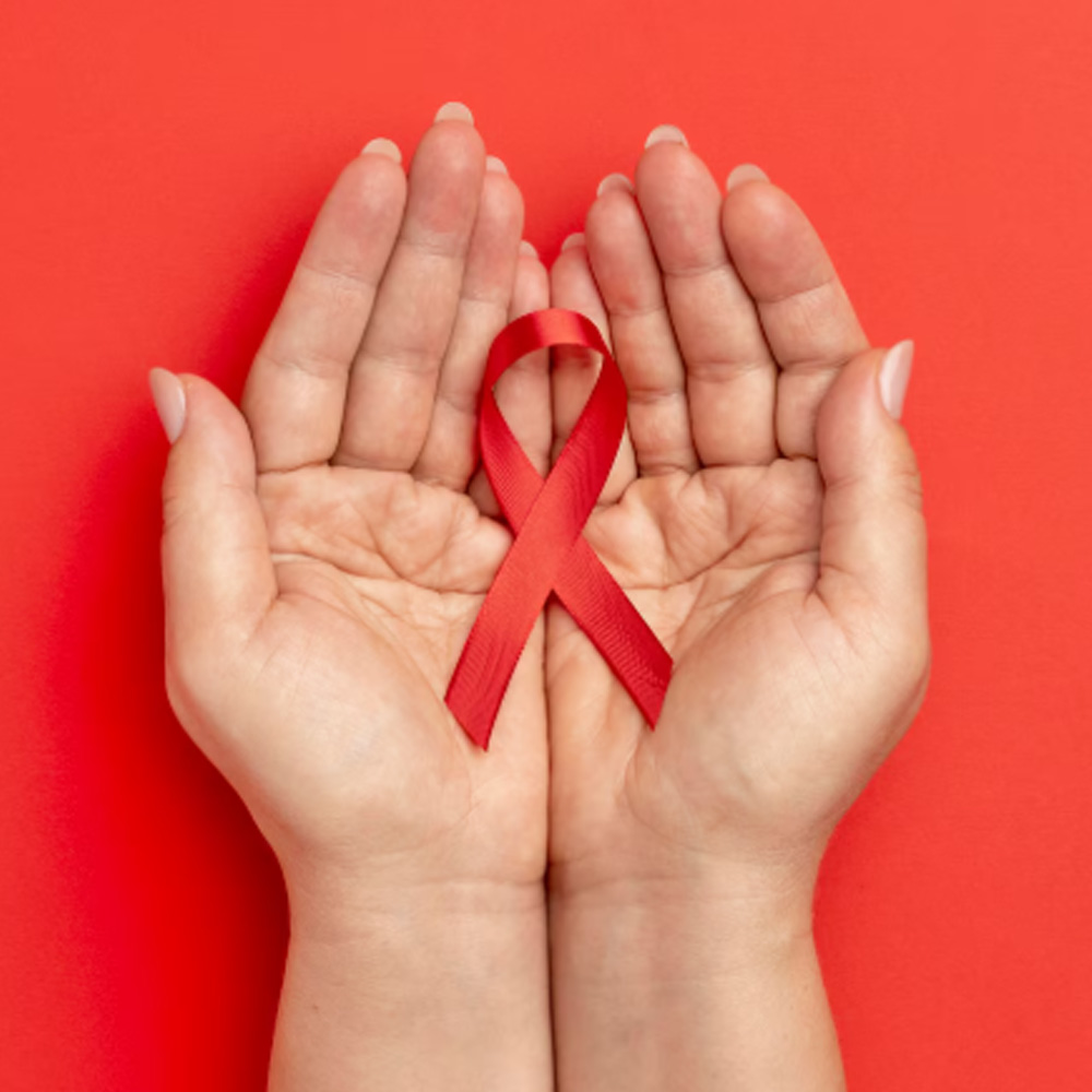 Na imagem, duas mãos seguram o laço vermelho, símbolo da campanha de luta, prevenção e conscientização sobre AIDS