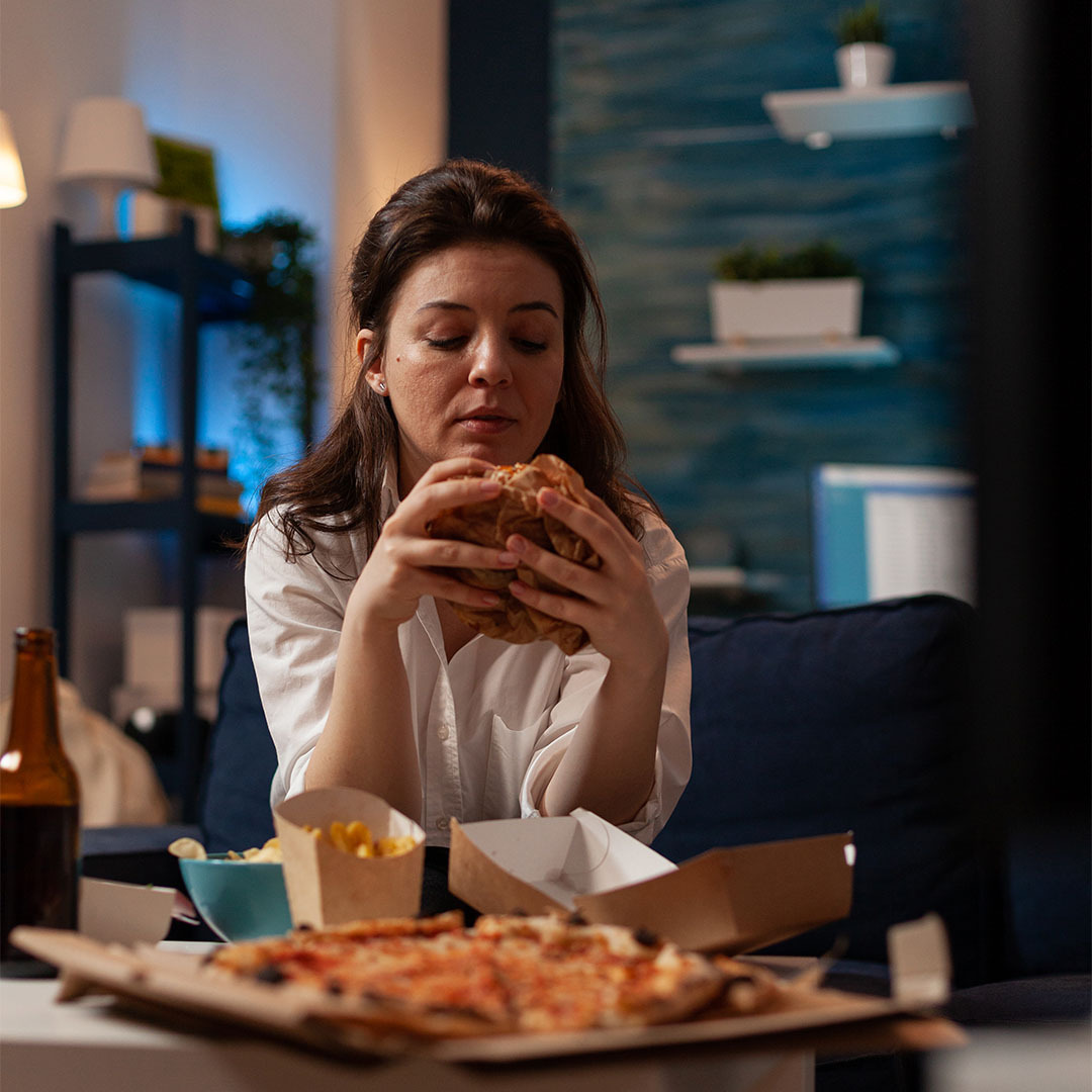 Na imagem, uma mulher sentada em uma poltrona comendo um hambúrguer e uma pizza em cima de uma mesa de centro à sua frente.