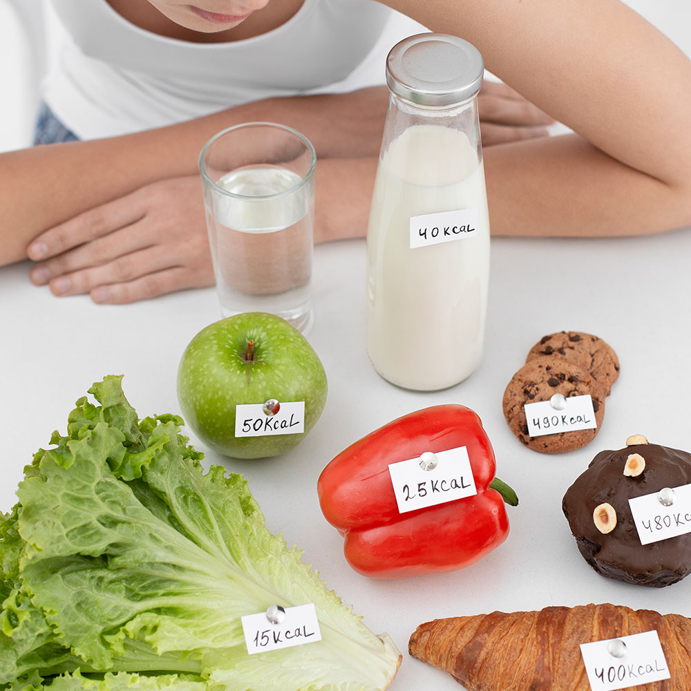 Na foto, alimentos com as suas respectivas calorias em uma mesa e uma pessoa apoiada na mesa observando.