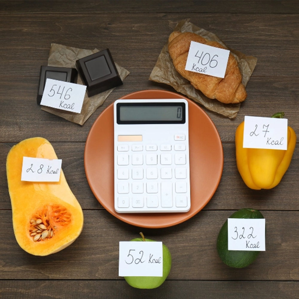 Na foto, uma calculadora em um prato rodeadas de alimentos contendo a quantidade específica de calorias.