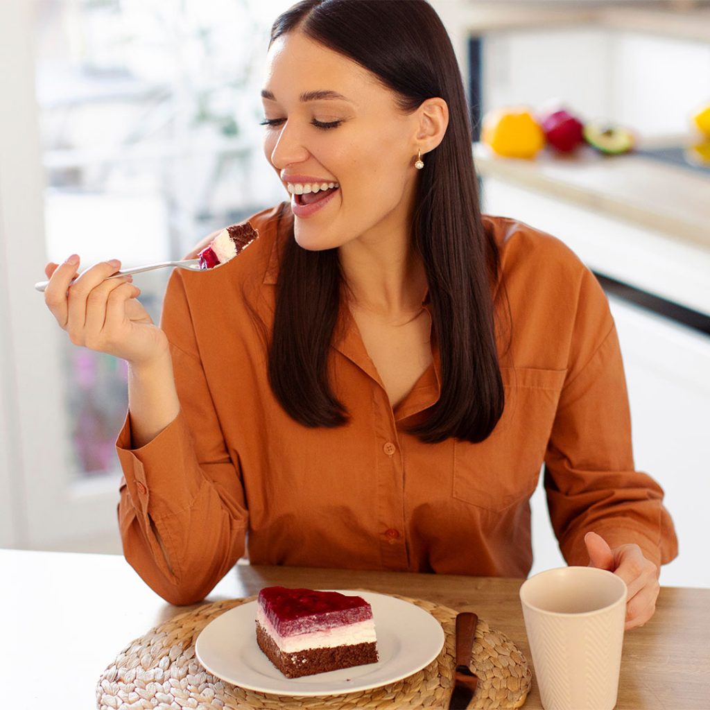 Mulher sorridente levando uma garfada de bolo de chocolate com creme e frutas vermelhas à boca, sentada à mesa da cozinha ao lado de uma caneca.