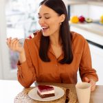 Mulher sorridente levando uma garfada de bolo de chocolate com creme e frutas vermelhas à boca, sentada à mesa da cozinha ao lado de uma caneca.