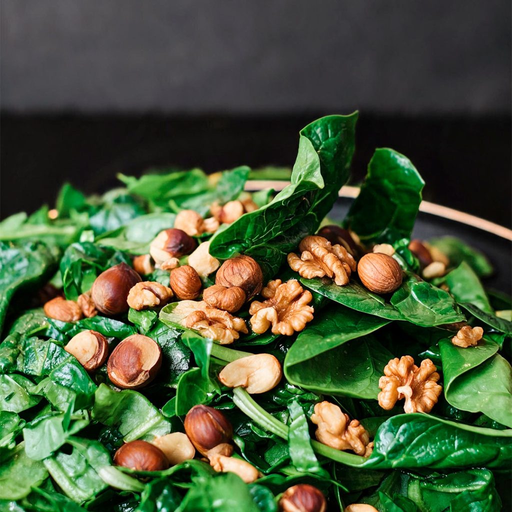 Salada de folhas verdes frescas coberta com nozes e avelãs inteiras em um prato sobre fundo escuro.