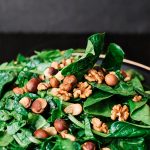 Salada de folhas verdes frescas coberta com nozes e avelãs inteiras em um prato sobre fundo escuro.