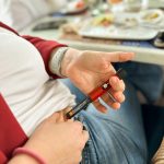 Pessoa aplicando medicação com uma caneta injetável vermelha no abdômen, sentada próximo a uma mesa de refeição.