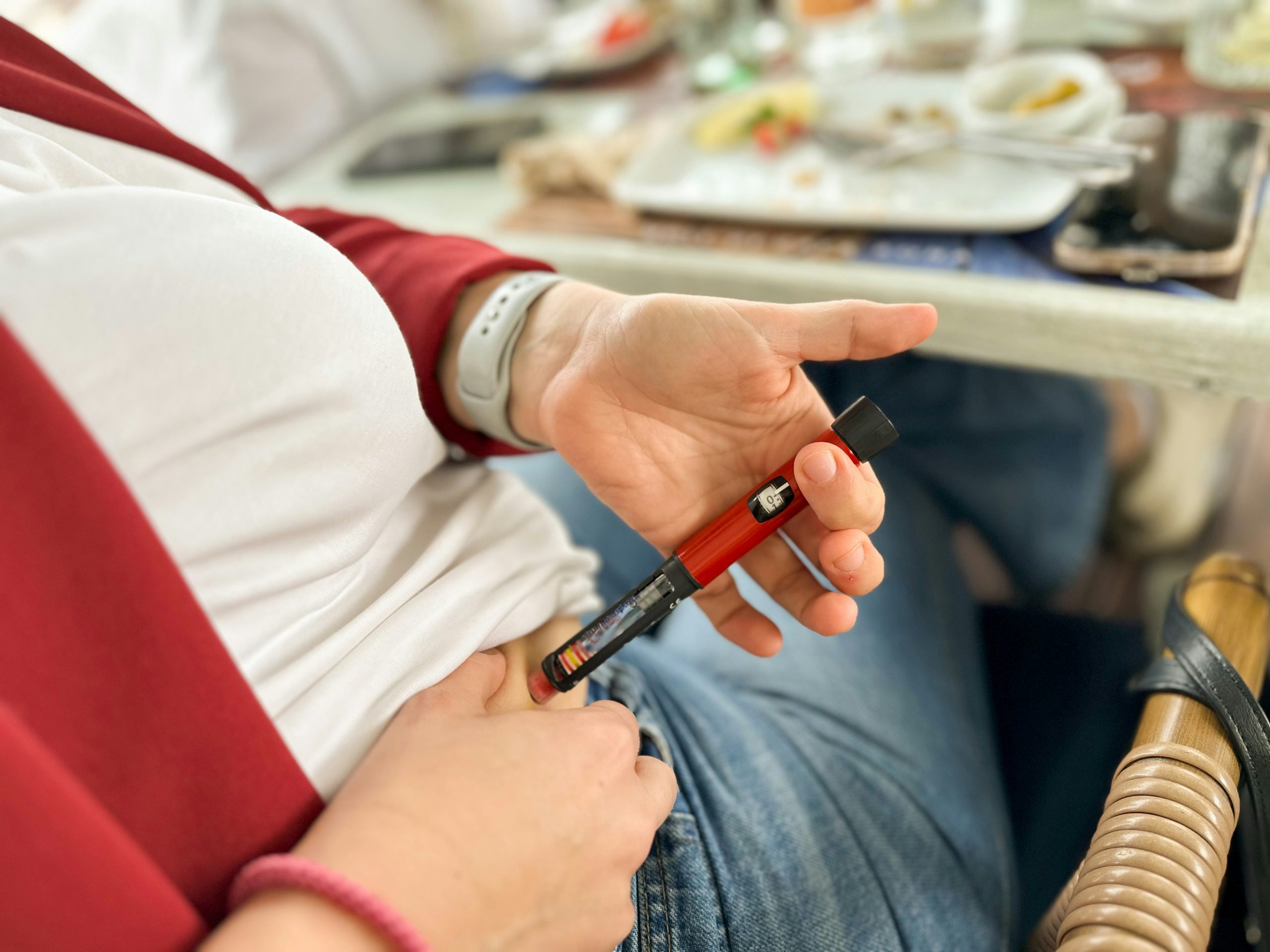 Pessoa aplicando medicação com uma caneta injetável vermelha no abdômen, sentada próximo a uma mesa de refeição.