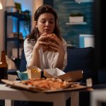 Mulher sentada em um sofá de sala à noite, segurando um hambúrguer grande com as duas mãos, diante de uma mesa repleta de fast-food com pizza, batatas fritas e salgadinhos.