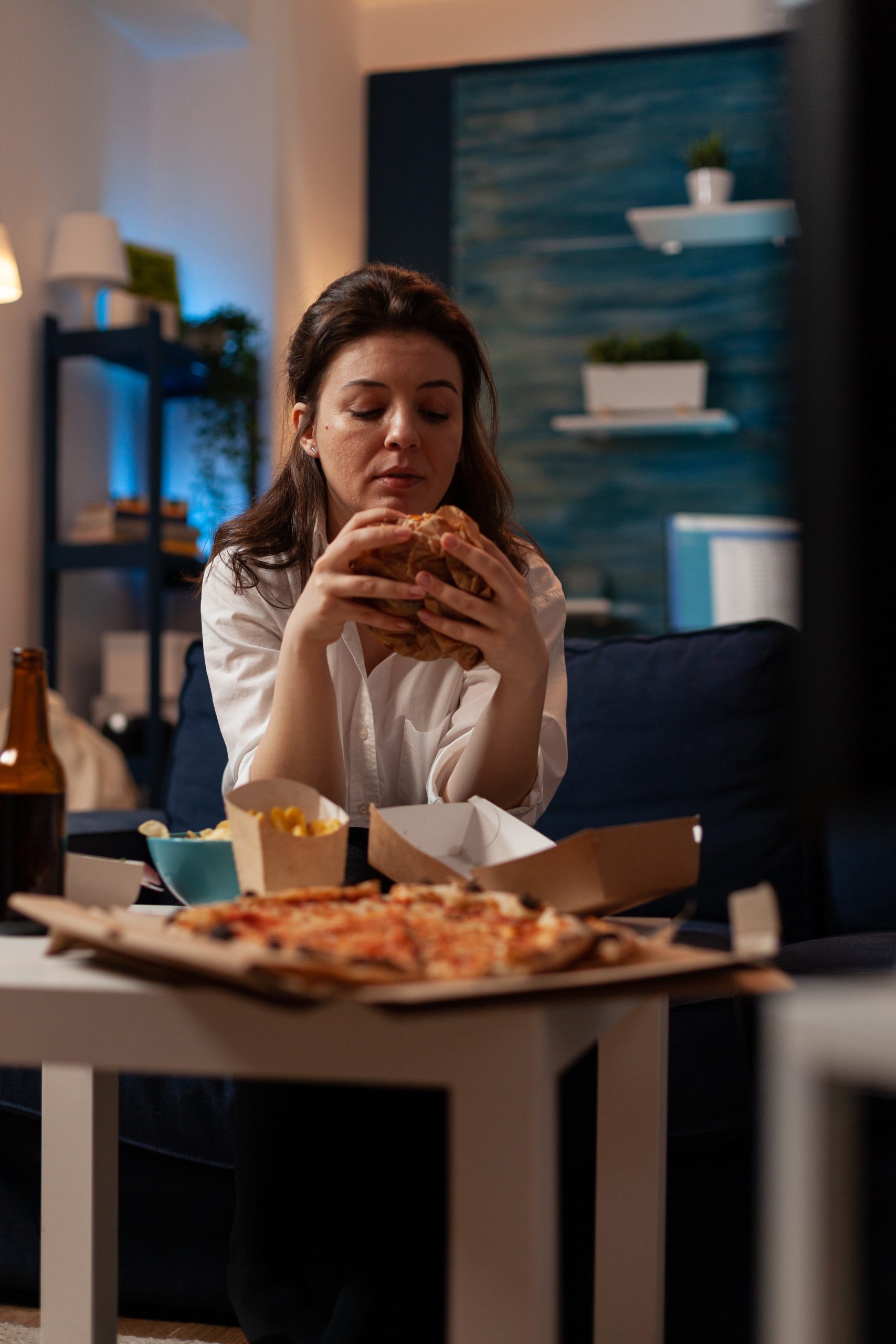 Mulher sentada em um sofá de sala à noite, segurando um hambúrguer grande com as duas mãos, diante de uma mesa repleta de fast-food com pizza, batatas fritas e salgadinhos.